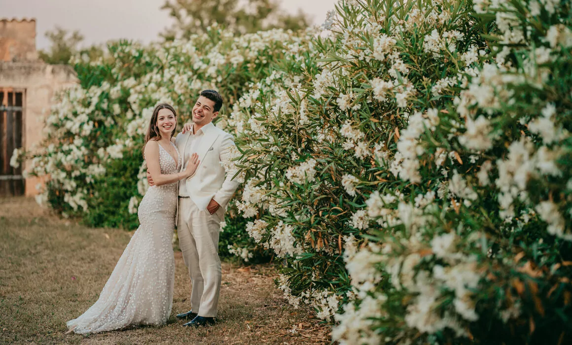 Eine Braut und ein Bräutigam stehen lächelnd zusammen im Freien neben hohen Büschen, die mit weißen Blumen bedeckt sind. Elegant gekleidet für ihre Hochzeit auf Mallorca, sehen sie strahlend aus - perfekt eingefangen von einem talentierten Hochzeitsfotografen Mallorca.