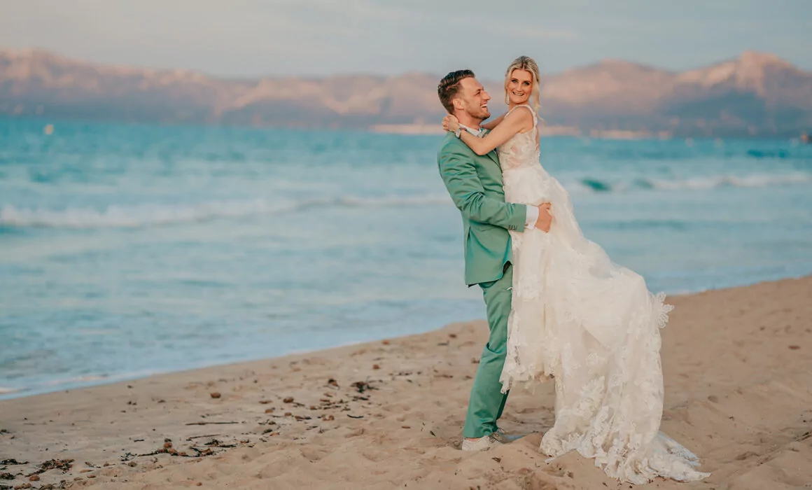 Ein Bräutigam in einem grünen Anzug hebt seine lächelnde Braut in einem weißen Spitzenkleid an einem Sandstrand, mit blauen Meereswellen und Bergen bei Sonnenuntergang - fangen Sie den Zauber einer Hochzeit auf Mallorca mit einem talentierten Hochzeitsfotografen Mallorca ein.