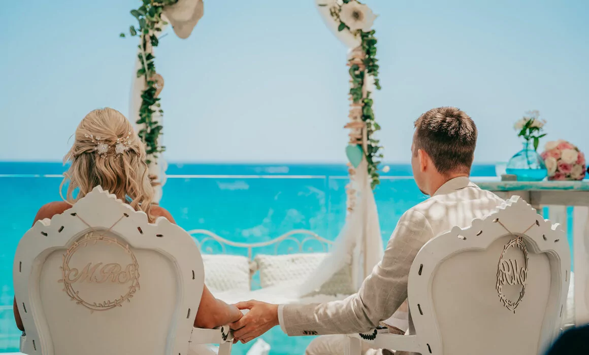 Braut und Bräutigam sitzen auf weißen, verzierten Stühlen, halten sich an den Händen und blicken auf einen geblümten Hochzeitsbogen mit dem Meer im Hintergrund - der perfekte Moment für einen Hochzeitsfotografen Mallorca, um Ihre unvergessliche Hochzeit auf Mallorca festzuhalten.