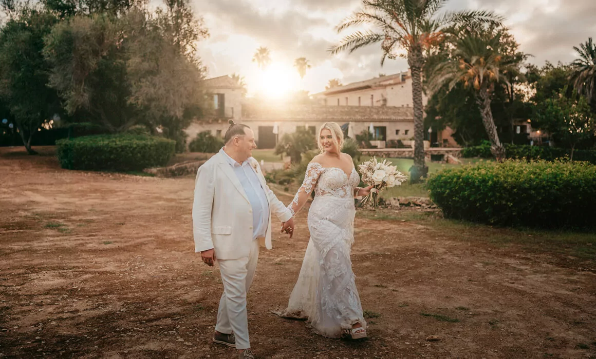 Eine Braut und ein Bräutigam in weißer Kleidung gehen bei Sonnenuntergang lächelnd Hand in Hand im Freien. Üppiges Grün, Palmen und ein rustikales Gebäude bilden die perfekte Kulisse für ihre Hochzeit auf Mallorca, während die Sonne warm hinter ihnen scheint.