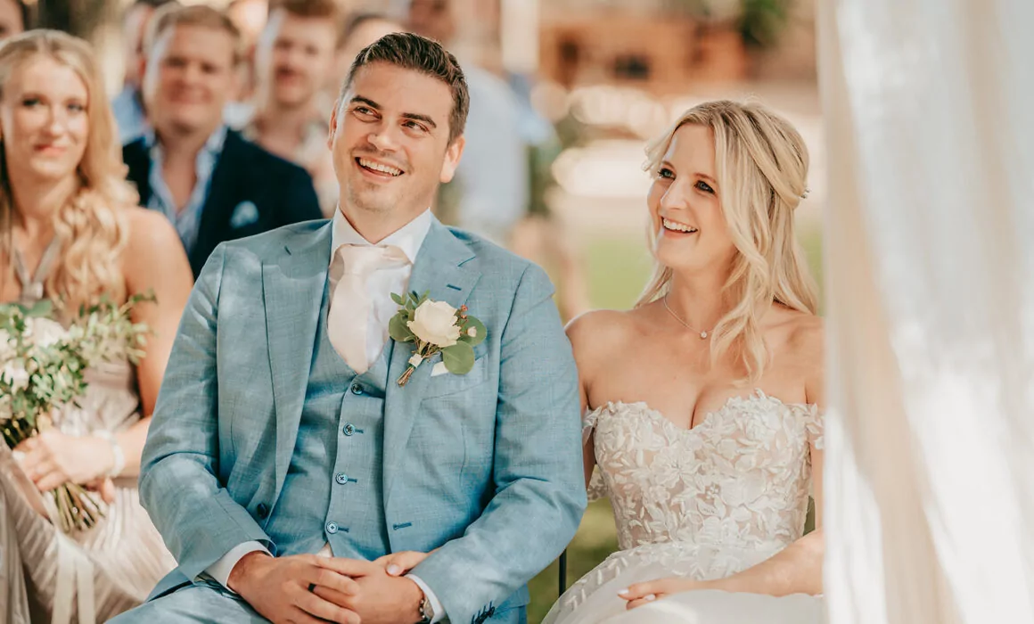 Eine Braut in einem weißen, trägerlosen Spitzenkleid und ein Bräutigam in einem hellblauen Anzug mit einer Boutonniere sitzen lächelnd nebeneinander bei ihrer Hochzeit auf Mallorca, mit Gästen, die hinter ihnen bei dieser schönen Hochzeitszeremonie im Freien sitzen.