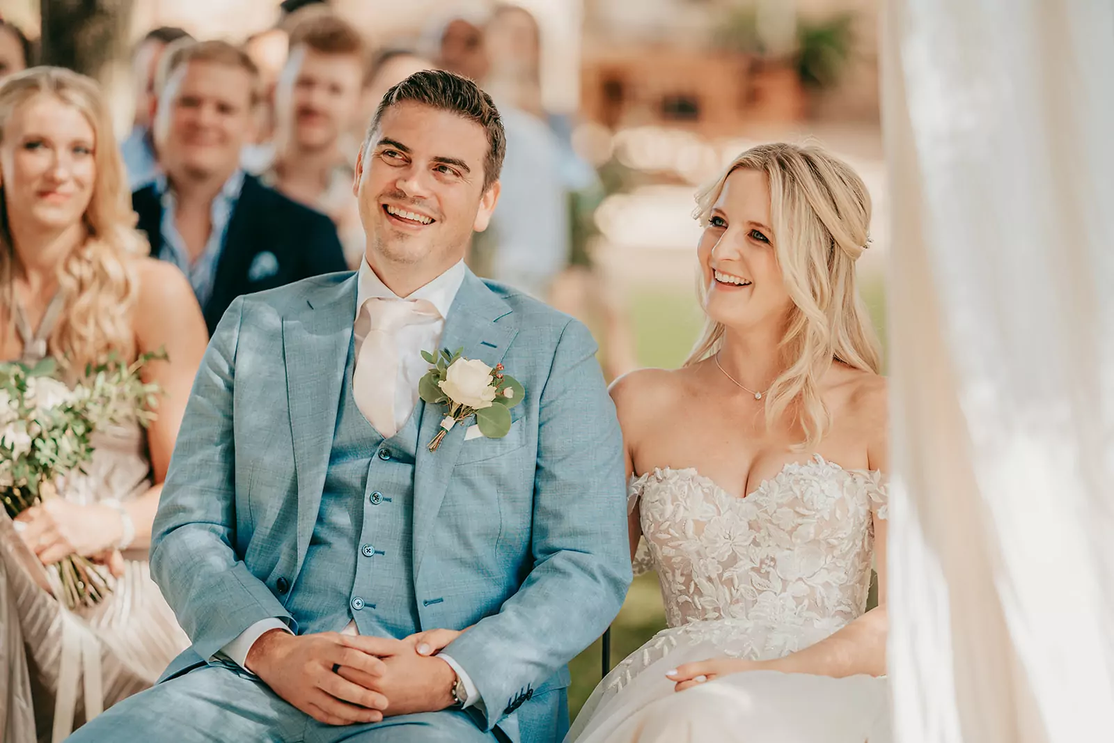 selina und sven: finca alaiar Eine Braut in einem weißen, trägerlosen Spitzenkleid und ein Bräutigam in einem hellblauen Anzug mit einer Boutonniere sitzen lächelnd nebeneinander bei ihrer Hochzeit auf Mallorca, mit Gästen, die hinter ihnen bei dieser schönen Hochzeitszeremonie im Freien sitzen.