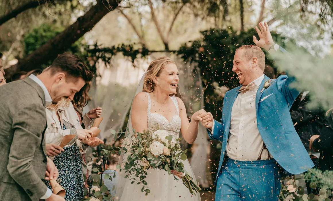 Ein fröhliches Brautpaar geht während ihrer Hochzeit auf Mallorca Hand in Hand im Freien spazieren, während die Gäste Konfetti werfen, um ihre Hochzeit zu feiern. Die Braut hält einen Blumenstrauß in der Hand und lächelt breit; der Bräutigam im blauen Anzug hebt aufgeregt seine Hand inmitten des üppigen Grüns.