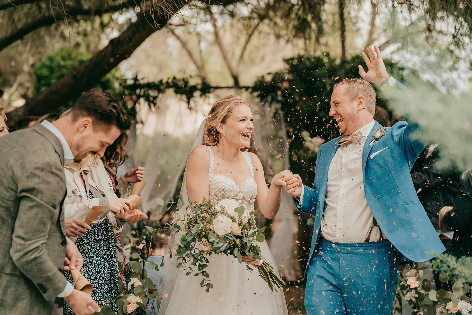 katja und benni: finca alaiar Ein fröhliches Brautpaar geht während ihrer Hochzeit auf Mallorca Hand in Hand im Freien spazieren, während die Gäste Konfetti werfen, um ihre Hochzeit zu feiern. Die Braut hält einen Blumenstrauß in der Hand und lächelt breit; der Bräutigam im blauen Anzug hebt aufgeregt seine Hand inmitten des üppigen Grüns.