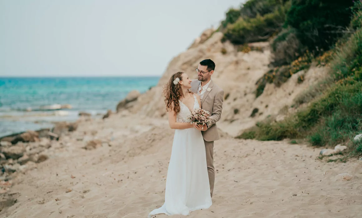 Eine Braut und ein Bräutigam stehen zusammen an einem Sandstrand, umarmen sich und lächeln sich an. Die Braut hält einen Blumenstrauß, mit dem Meer und den felsigen Klippen im Hintergrund - ein perfekter Moment für jeden Hochzeitsfotograf Mallorca bei einer Hochzeit auf Mallorca.