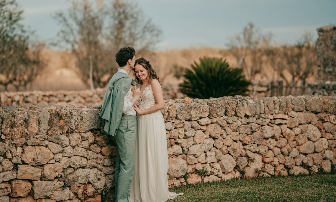 Ein Paar in Hochzeitskleidung steht an einer rustikalen Steinmauer auf der Finca Alaiar. Der Mann küsst sanft die Stirn der Frau, während sie lächelnd seinen Arm hält. Bäume und sanftes Abendlicht vervollständigen diese romantische Hochzeitsfotograf Mallorca Szene.