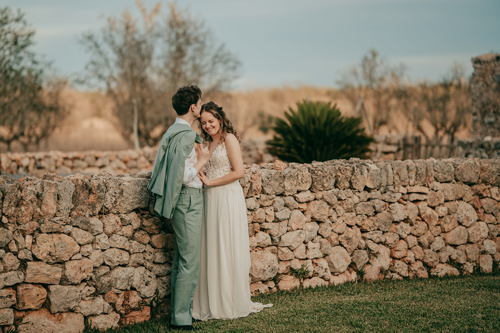 Hochzeit auf Finca Alaiar Mallorca Ein Paar in Hochzeitskleidung steht an einer rustikalen Steinmauer auf der Finca Alaiar. Der Mann küsst sanft die Stirn der Frau, während sie lächelnd seinen Arm hält. Bäume und sanftes Abendlicht vervollständigen diese romantische Hochzeitsfotograf Mallorca Szene.