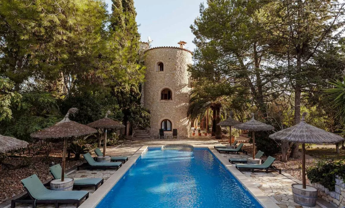Eine Villa im Stil eines Steinturms in Can Davero, umgeben von Bäumen, mit einem rechteckigen Swimmingpool im Vordergrund, der von Sonnenliegen und strohgedeckten Sonnenschirmen umgeben ist - ein idyllischer Ort für jeden Hochzeitsfotografen Mallorca.