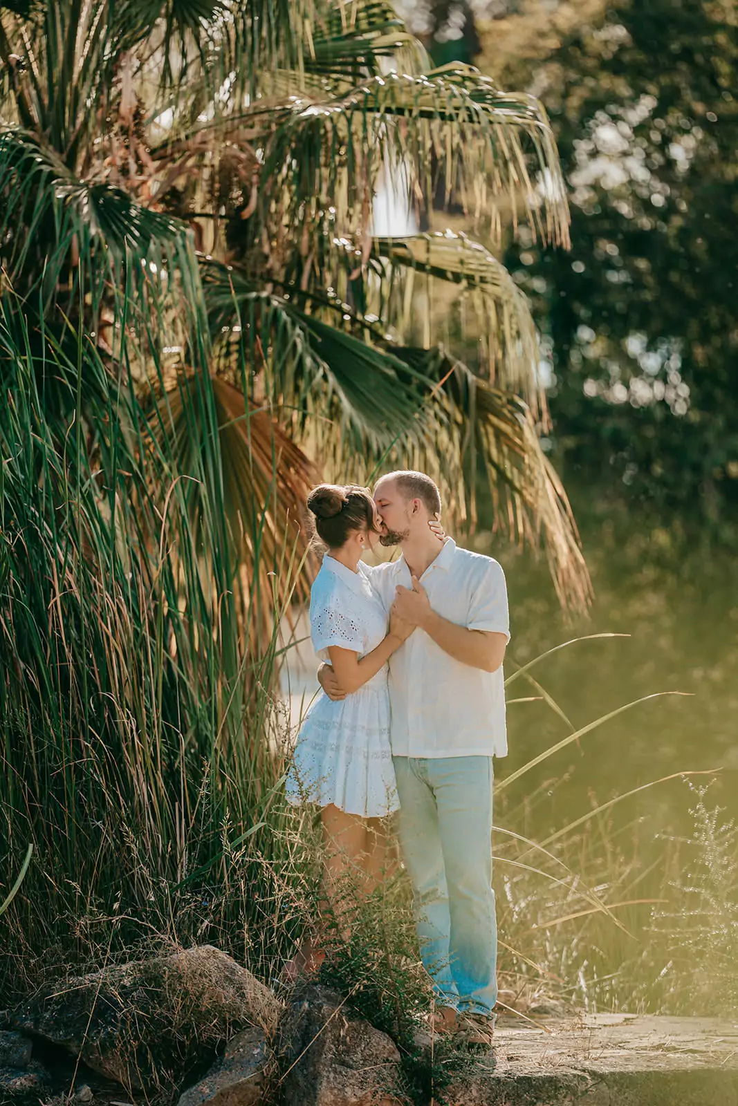 Ein Paar steht im Freien zwischen hohem Gras und Palmen in Can Davero und küsst sich innig. Bekleidet mit heller Sommerkleidung, sonnen sie sich im Sonnenlicht - ein perfekter Moment, eingefangen von einem Hochzeitsfotografen Mallorca.