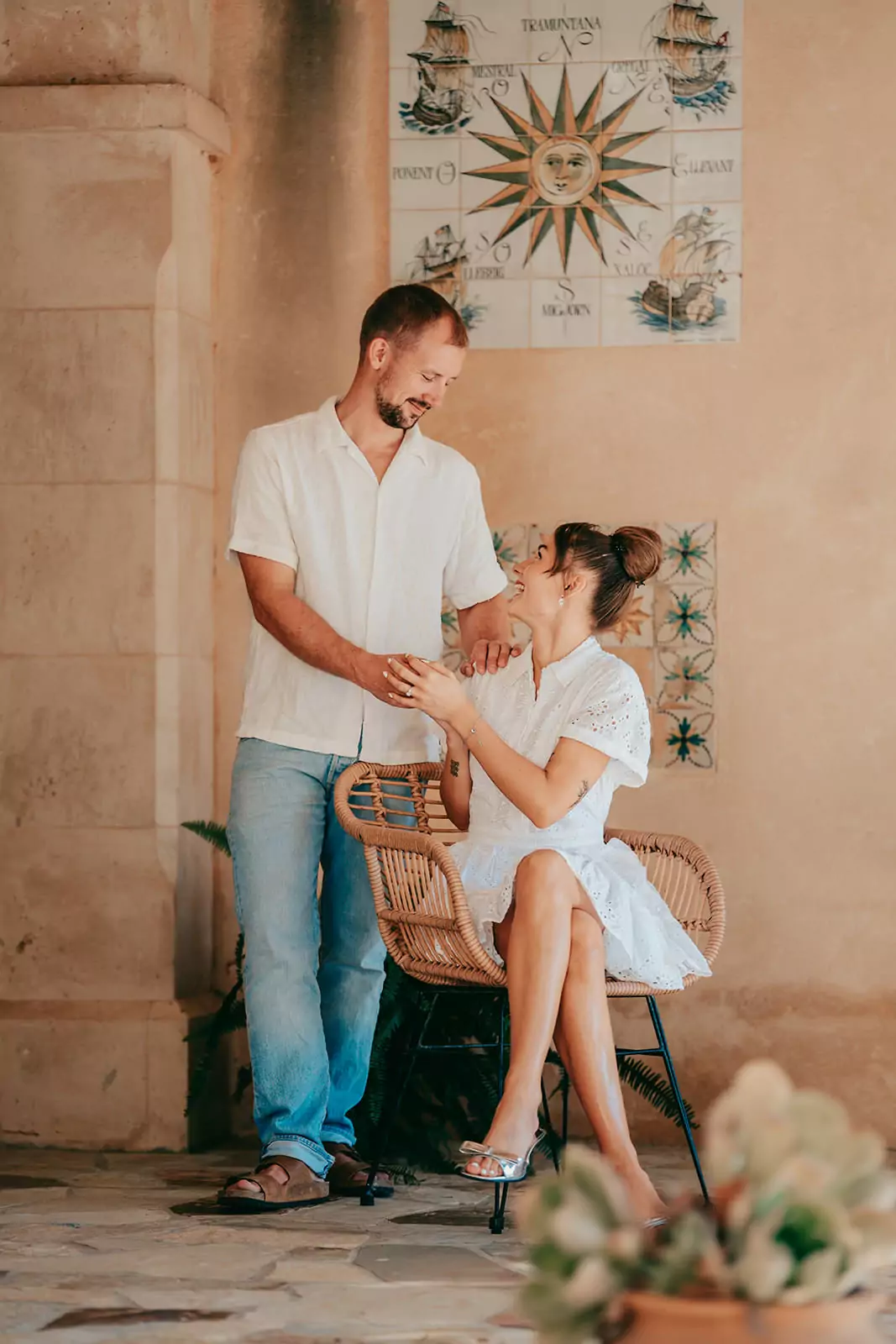 Ein Mann in einem weißen Hemd und Jeans steht neben einer sitzenden Frau in einem weißen Kleid in Can Davero. Sie lächeln sich an und halten sich an den Händen, während Hochzeitsfotograf Mallorca ihren Moment in einem rustikalen, sonnenbeschienenen Innenhof mit Steinmauern und dekorativen Fliesen festhält.