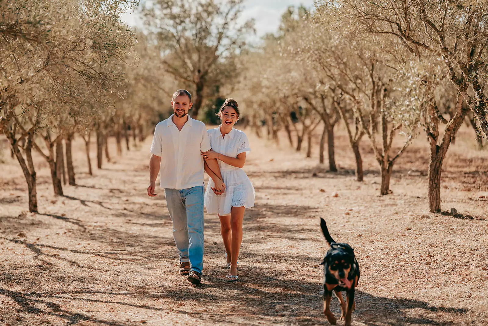Ein lächelndes Paar spaziert Arm in Arm über einen sonnenbeschienenen, von Bäumen gesäumten Feldweg in Can Davero, während ein schwarz-brauner Hund vorausläuft. Die Szene sieht warm und entspannt aus, mit trockenen Blättern auf dem Boden - perfekt für eine Hochzeitsfotograf Mallorca-Session.