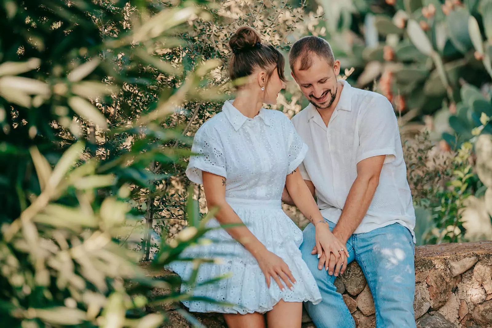 Ein Paar sitzt auf einem Steinvorsprung in Can Davero, hält sich an den Händen und lächelt sich an. Umgeben von Grün, in ihren leichten, sommerlichen Outfits, verkörpern sie die warmen und entspannten Momente, die ein Hochzeitsfotograf Mallorca eingefangen hat.