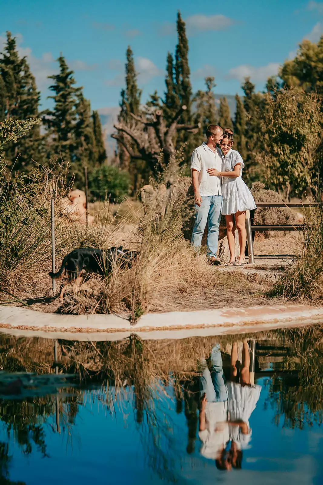 Ein Paar steht eng beieinander an einem Teich in einer sonnigen, natürlichen Umgebung mit hohen Bäumen im Hintergrund und ihrem Hund in der Nähe. Aufgenommen von Hochzeitsfotograf Mallorca Can Davero, schimmert ihr Spiegelbild sanft im Wasser.