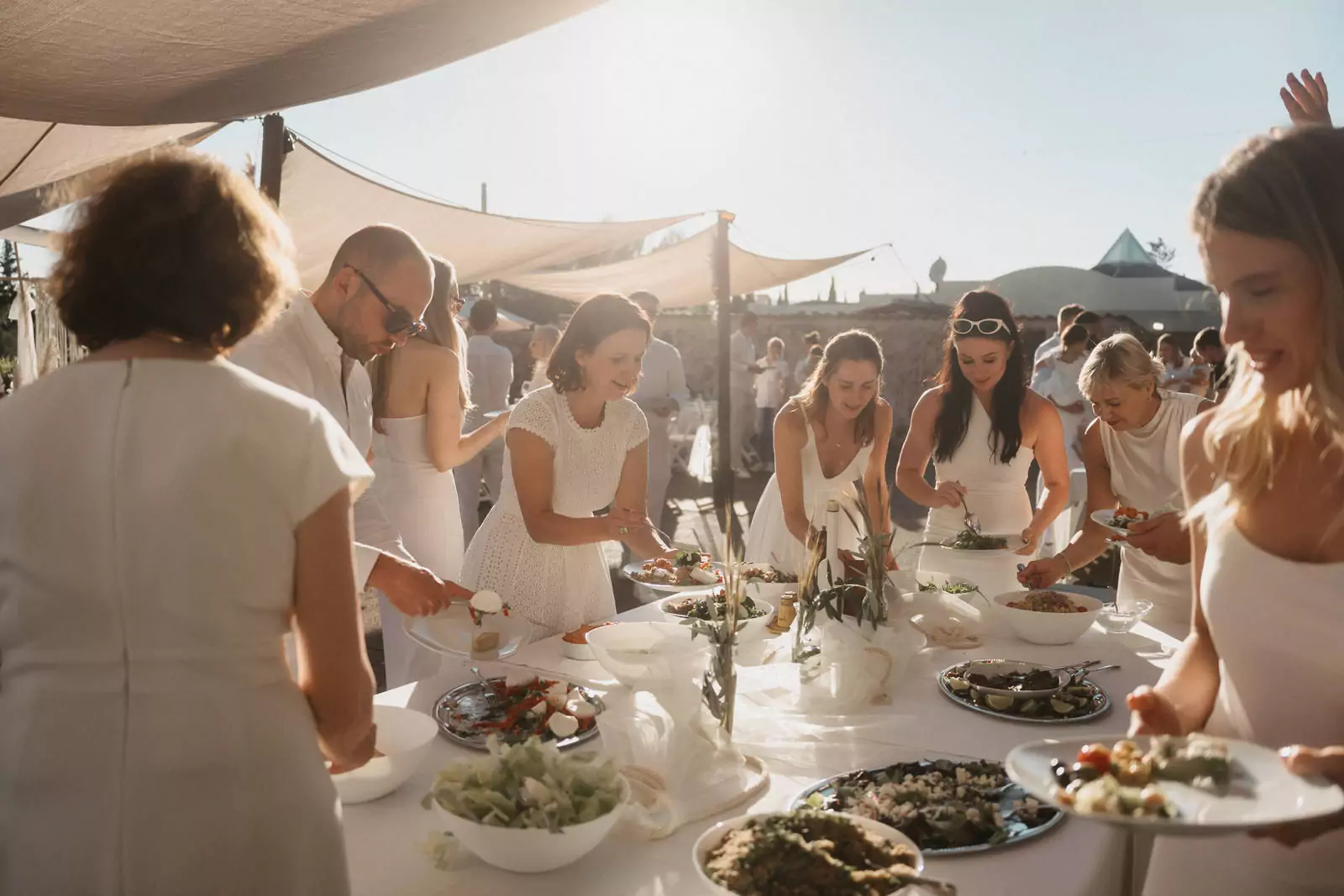 Eine Gruppe von weiß gekleideten Menschen bedient sich bei einer Veranstaltung im Freien in Can Davero an einem Buffet, während die Sonne über die Szene fällt. Der Tisch ist mit verschiedenen Gerichten und Salaten gedeckt, aufgenommen von einem Hochzeitsfotograf Mallorca.