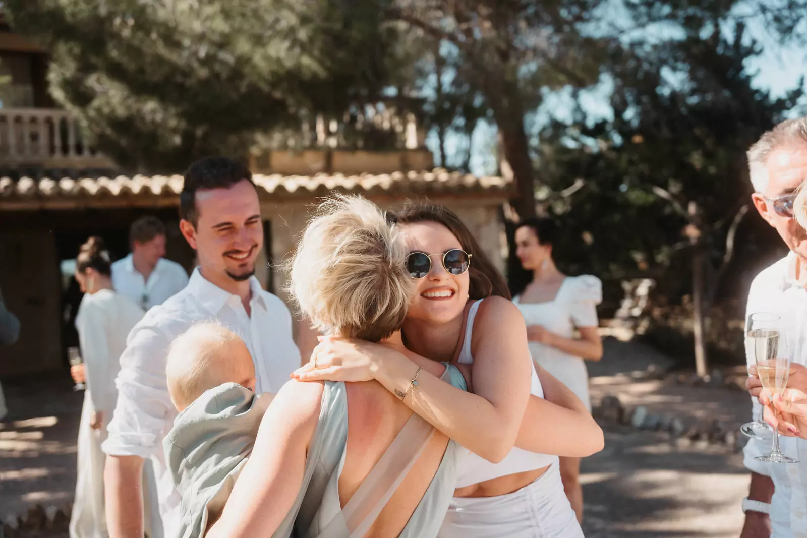 In Can Davero umarmt eine Frau mit Sonnenbrille eine andere Frau, die ein Baby hält, bei einem Treffen im Freien. Andere Gäste lächeln in der Nähe, Getränke in der Hand, während der Hochzeitsfotograf Mallorca Erinnerungen vor rustikalen Gebäuden und Bäumen festhält.