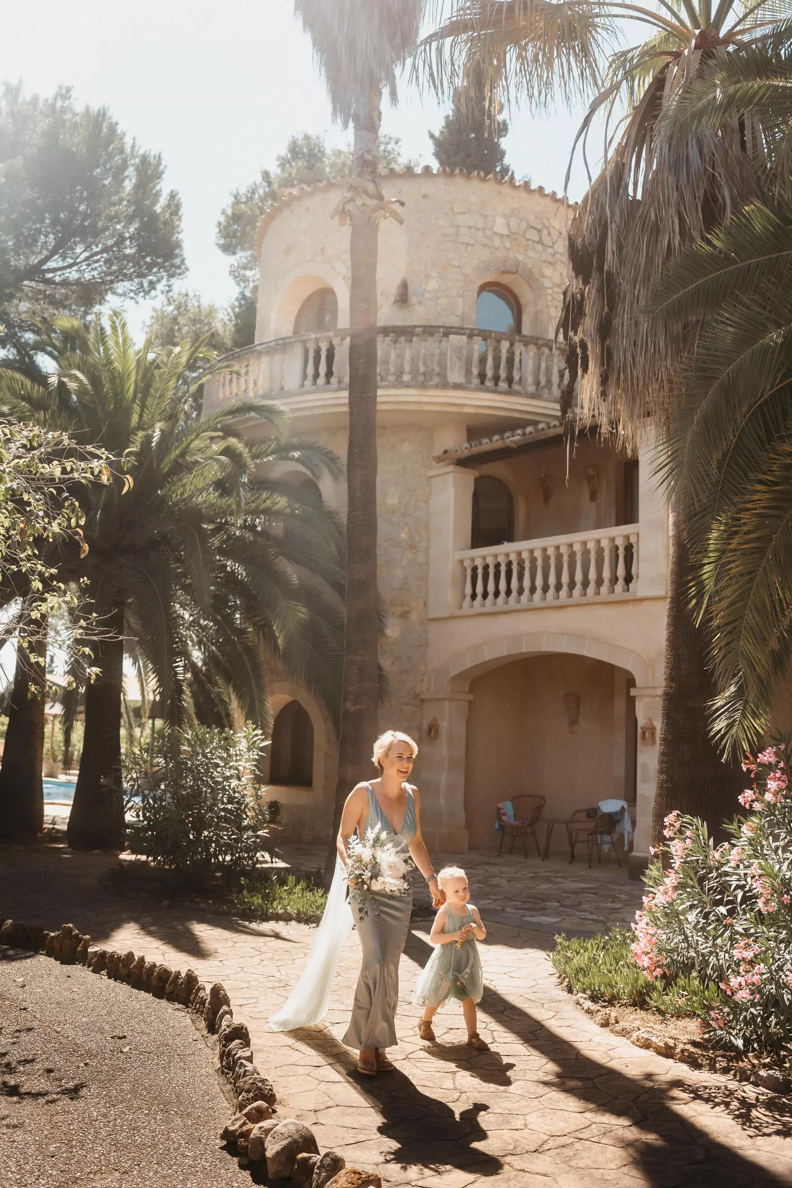 Eine Frau in einem hellen Kleid, die einen Blumenstrauß in der Hand hält, geht Hand in Hand mit einem kleinen Kind einen sonnenbeschienenen Weg in Can Davero entlang, mit Palmen und einer rustikalen Villa im mediterranen Stil im Hintergrund - aufgenommen von Hochzeitsfotograf Mallorca.