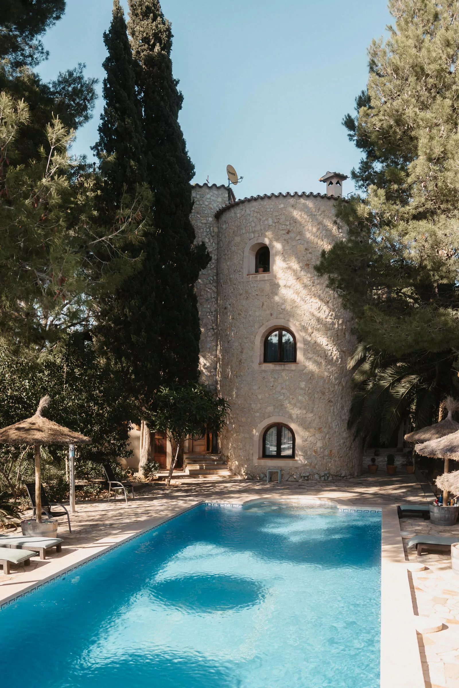 Eine Steinvilla in Can Davero mit gewölbten Fenstern, umgeben von hohen Bäumen, steht hinter einem klaren blauen Pool. Sonnenliegen und strohgedeckte Sonnenschirme säumen die Terrasse - ein idealer Rahmen für einen Hochzeitsfotografen Mallorca, um perfekte Momente einzufangen.