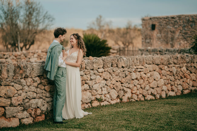 Ein Paar in Hochzeitskleidung lächelt sich an einer rustikalen Steinmauer an, umgeben von Bäumen und Steingebäuden auf der Finca Alaiar - eine ideale Szene für jeden Hochzeitsfotografen Mallorca, um zeitlose Momente einzufangen.