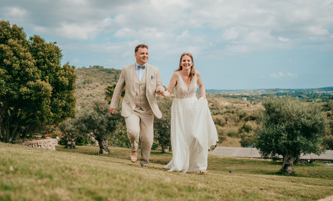 Braut und Bräutigam in Hochzeitskleidung gehen freudig Hand in Hand einen grasbewachsenen Hügel auf der Finca Ses Voltes hinauf, mit Bäumen und einer malerischen Landschaft im Hintergrund unter einem teilweise bewölkten Himmel.