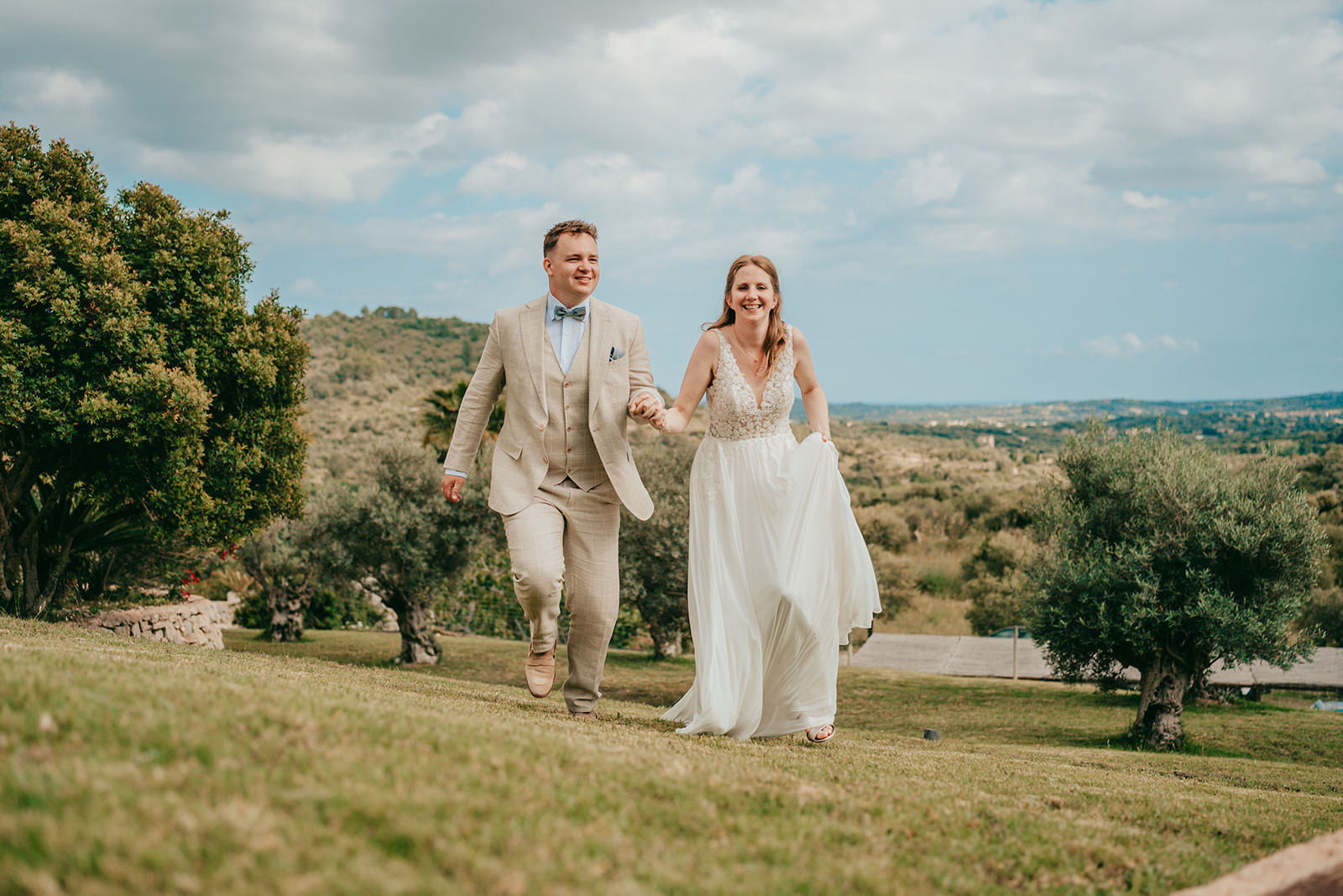 A smiling bride and groom hold hands and walk up a grassy hill at Finca Ses Voltes, surrounded by trees and a scenic landscape under a partly cloudy sky—an unforgettable Ses Voltes Hochzeit moment.
