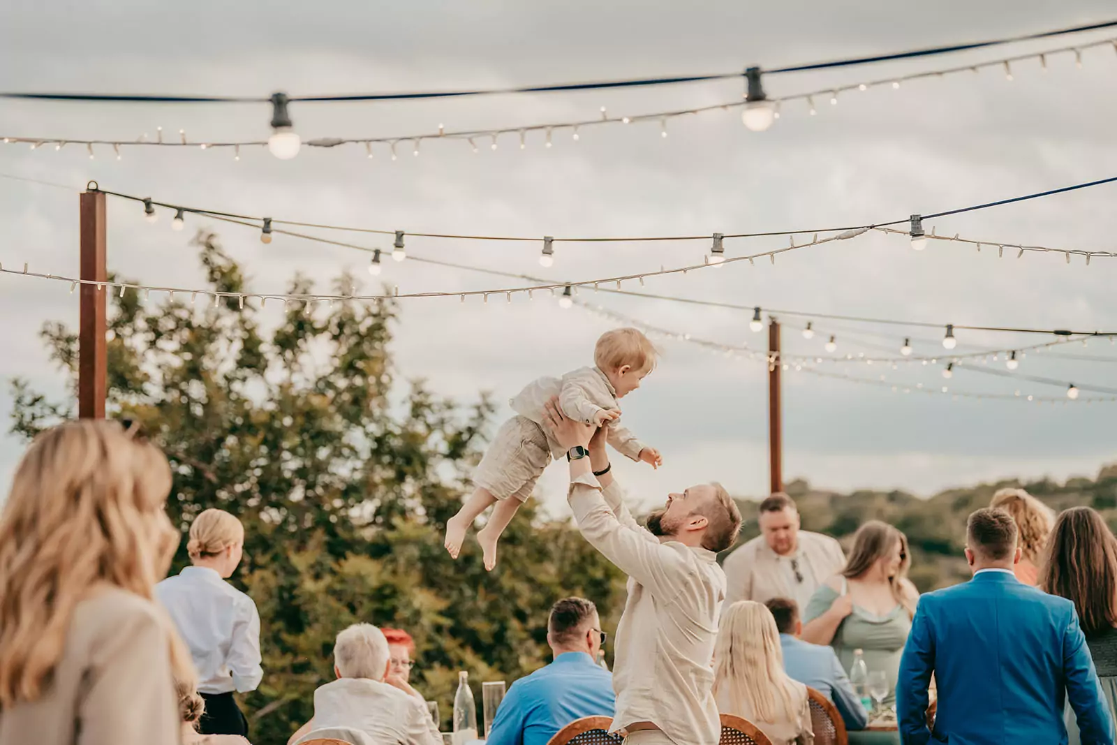 Ein Mann hebt ein lächelndes Kleinkind in die Luft bei einer fröhlichen Versammlung im Freien auf der Finca Ses Voltes. Die Menschen, die eine Hochzeit auf Mallorca feiern, sitzen und stehen unter Lichterketten mit Bäumen und einem bewölkten Himmel im Hintergrund.