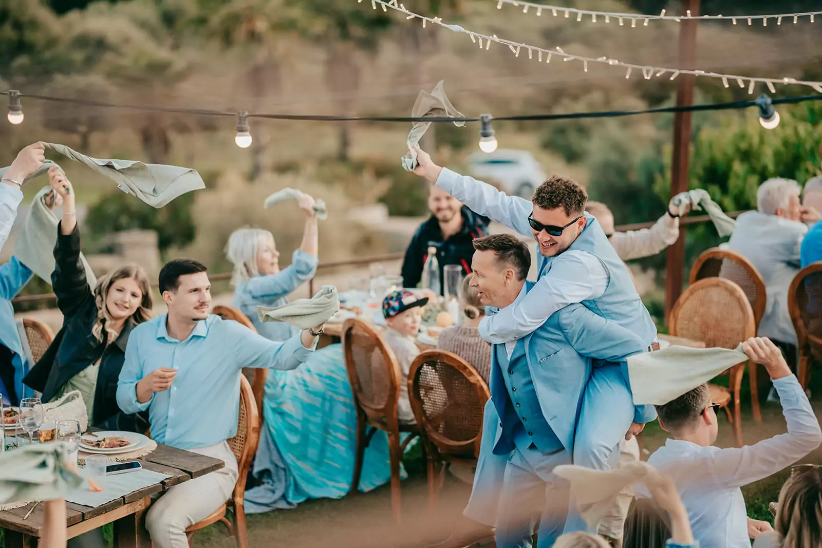 Bei einer Ses-Voltes-Mallorca-Hochzeit winken die Gäste fröhlich mit Servietten, während ein Mann im blauen Anzug und mit Sonnenbrille auf einem anderen Mann Huckepack reitet. An den festlich geschmückten Tischen unter den leuchtenden Lichterketten wird viel gelacht.