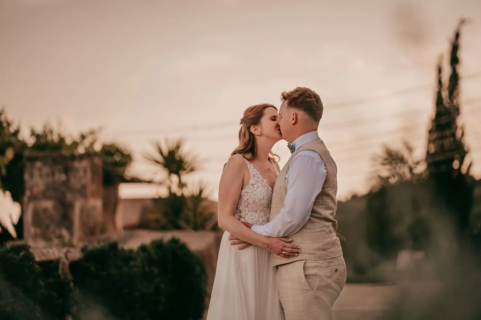 Ein Paar in Hochzeitskleidung umarmt und küsst sich im Freien bei Sonnenuntergang in Ses Voltes Mallorca, umgeben von Grün und weichem, warmem Licht - perfekt für eine romantische Hochzeit.
