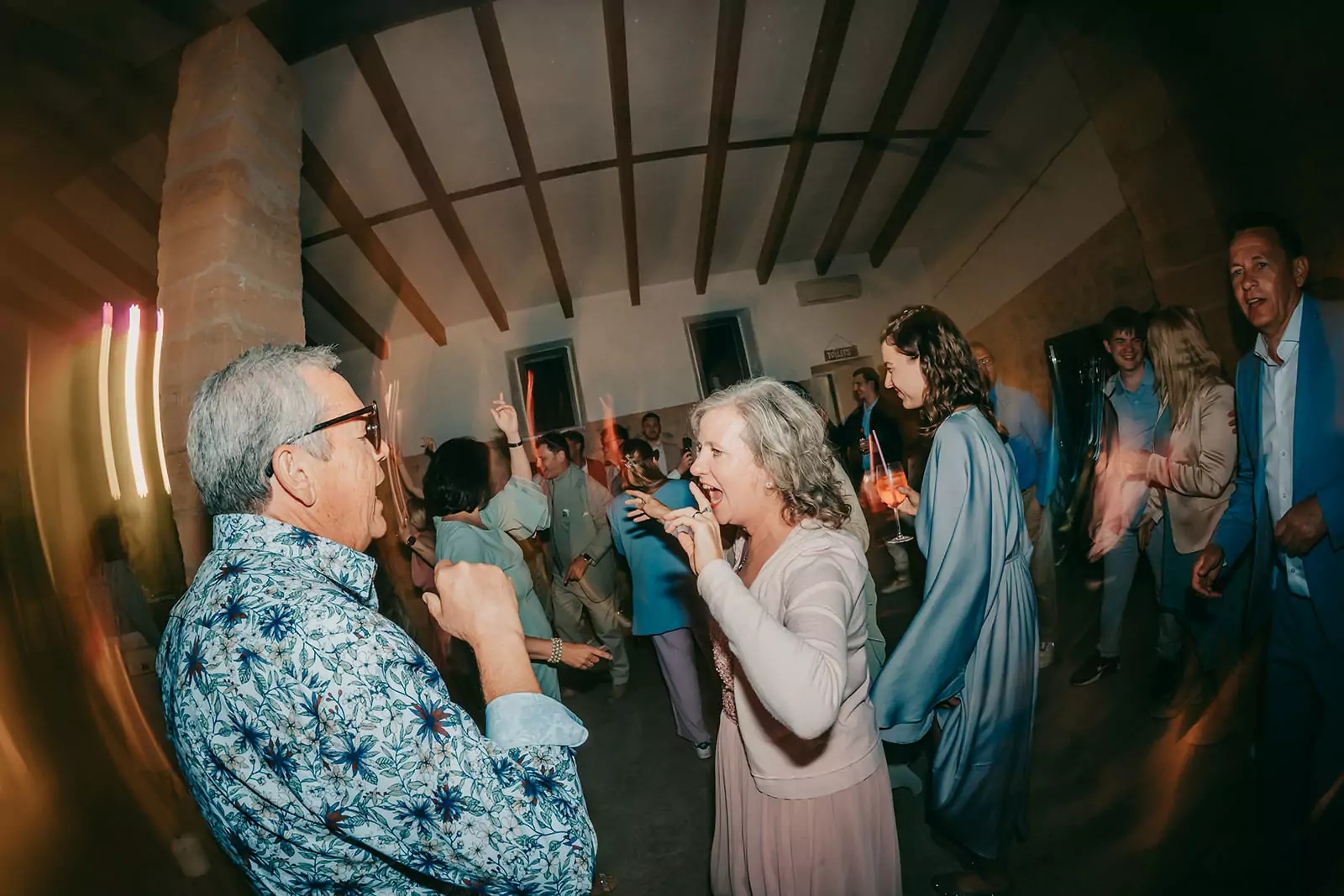 Eine lebhafte Gruppe von Menschen, meist Erwachsene, tanzt und lacht zusammen in einem warm beleuchteten Innenraum mit Holzbalken an der Decke im Ses Voltes Mallorca. Die festliche Atmosphäre ist perfekt für eine fröhliche Hochzeitsfeier.