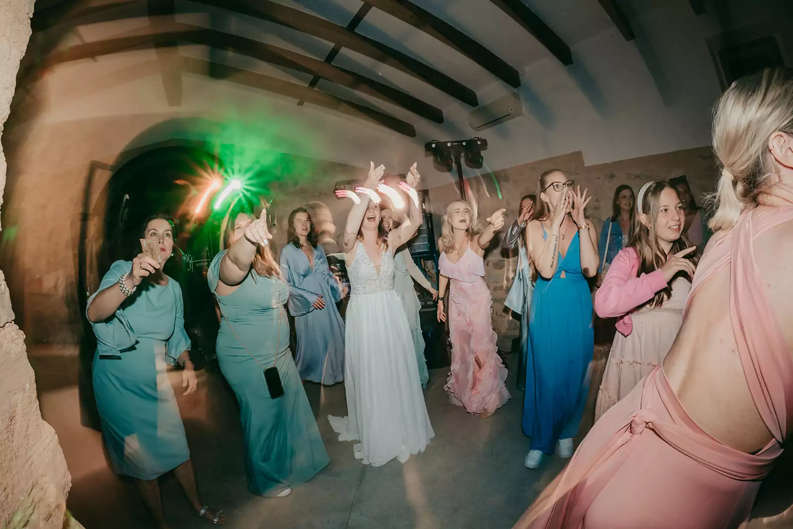 Eine Gruppe von Frauen in farbenfrohen Kleidern tanzt und feiert fröhlich unter grünen und rosafarbenen Lichtern auf einer Ses-Voltes-Mallorca-Hochzeit, wobei eine Frau in einem weißen Kleid in der Mitte die Arme hebt.