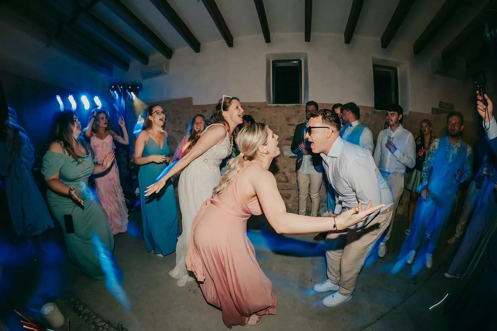 Eine Gruppe von Menschen in formeller Kleidung tanzt begeistert bei einer Indoor ses voltes mallorca hochzeit. Zwei Gäste, eine Frau in einem rosa Kleid und ein Mann in einem weißen Hemd, stehen sich gegenüber und lachen, während die Menge um sie herum jubelt. Blaue Lichter erhellen die Szene.