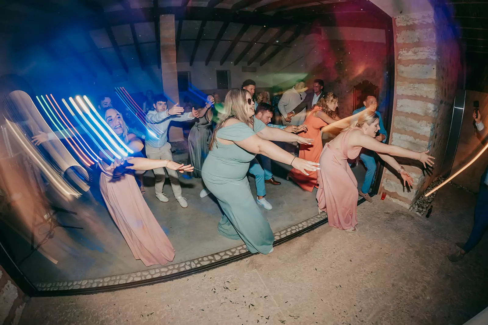 Eine Gruppe von Menschen in farbenfrohen Kleidern und Anzügen tanzt enthusiastisch mit ausgestreckten Armen, lächelnd und lachend, unter leuchtenden Partylichtern auf einer rustikalen Ses Voltes Mallorca Hochzeitsfeier.