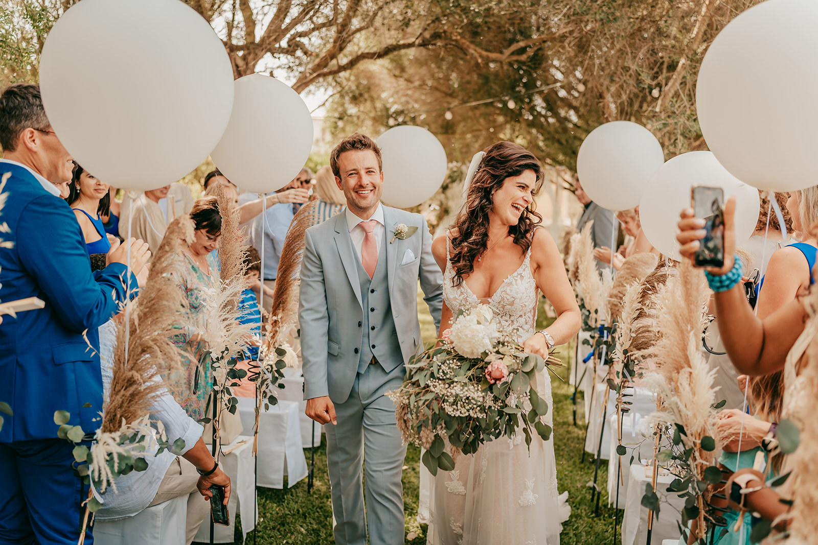 Hochzeit auf Finca Alaiar Mallorca Eine lächelnde Braut und ein lächelnder Bräutigam schreiten einen Gang im Freien auf der Finca Alaiar hinunter, umgeben von Gästen mit weißen Luftballons und Pampasgras unter einem Baldachin von Bäumen, wunderschön eingefangen von einem Hochzeitsfotograf Mallorca.
