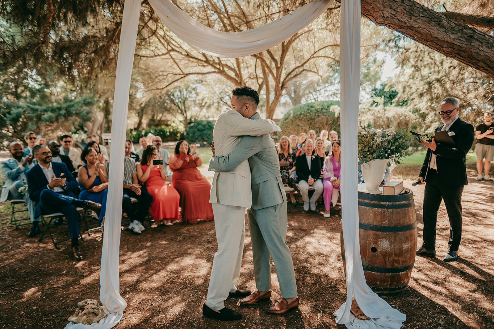 Hochzeit auf Finca Alaiar Mallorca Zwei Bräutigame in hellen Anzügen umarmen sich unter einem geschmückten Bogen bei ihrer Hochzeit im Freien auf der Finca Alaiar, umgeben von jubelnden Gästen zwischen Bäumen, mit einem Mann, der in der Nähe neben einem Holzfass die Trauung vollzieht - aufgenommen von einem talentierten Hochzeitsfotografen Mallorca.
