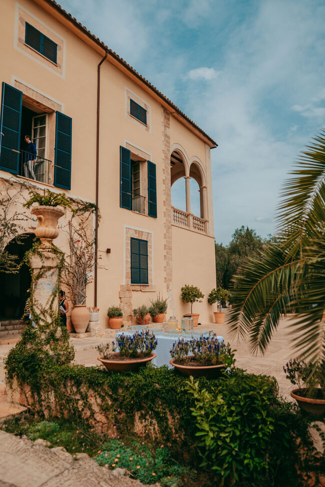 A Mediterranean-style villa with yellow walls, blue shutters, arched balcony, potted plants, and lush greenery under a blue sky. A person stands on a balcony, partially visible through an open door.