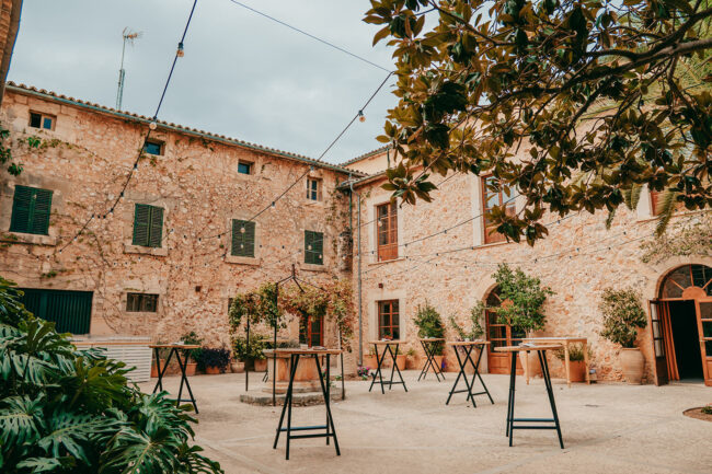 A rustic stone courtyard with string lights overhead, tall bar tables with no chairs, potted plants, and green shutters on the windows; a relaxed, outdoor gathering space.