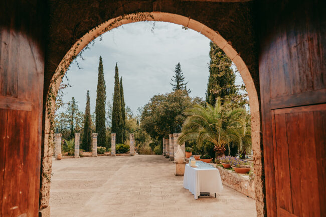 View through a large stone archway with wooden doors, showing a sunny patio with potted plants, tall cypress trees, and a white table covered with a cloth, set amid lush greenery under a cloudy sky.