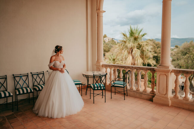 A bride in a white gown stands on a tiled terrace with columns, looking out at palm trees and distant hills. Several black chairs and a small round table are arranged nearby.