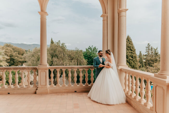 A bride in a white gown and a groom in a suit stand together on a grand balcony with columns, surrounded by lush greenery and trees under a cloudy sky.