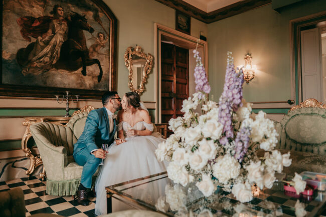 A bride and groom sit on an ornate sofa, sharing a kiss in an elegant, vintage room with classic decor, gold accents, and a large bouquet of white and purple flowers on a glass table in the foreground.