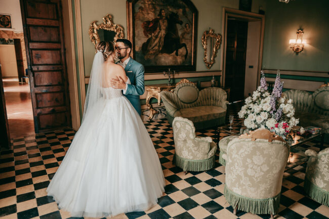 A bride and groom share a dance in an elegant, vintage room with checkered floors, antique furniture, and ornate gold-framed mirrors and paintings. The bride wears a white gown; the groom is in a light blue suit.