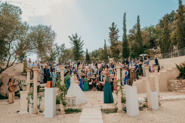 A bride and groom stand at an outdoor wedding ceremony, surrounded by bridesmaids in teal dresses and a large group of guests seated on stone steps, with greenery and columns decorating the venue under a sunny sky.