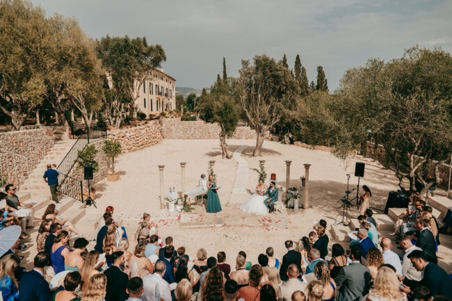 An outdoor wedding ceremony with two brides standing at the altar, surrounded by guests seated in rows. The setting is rustic, with stone structures, trees, and a sunny, open courtyard.