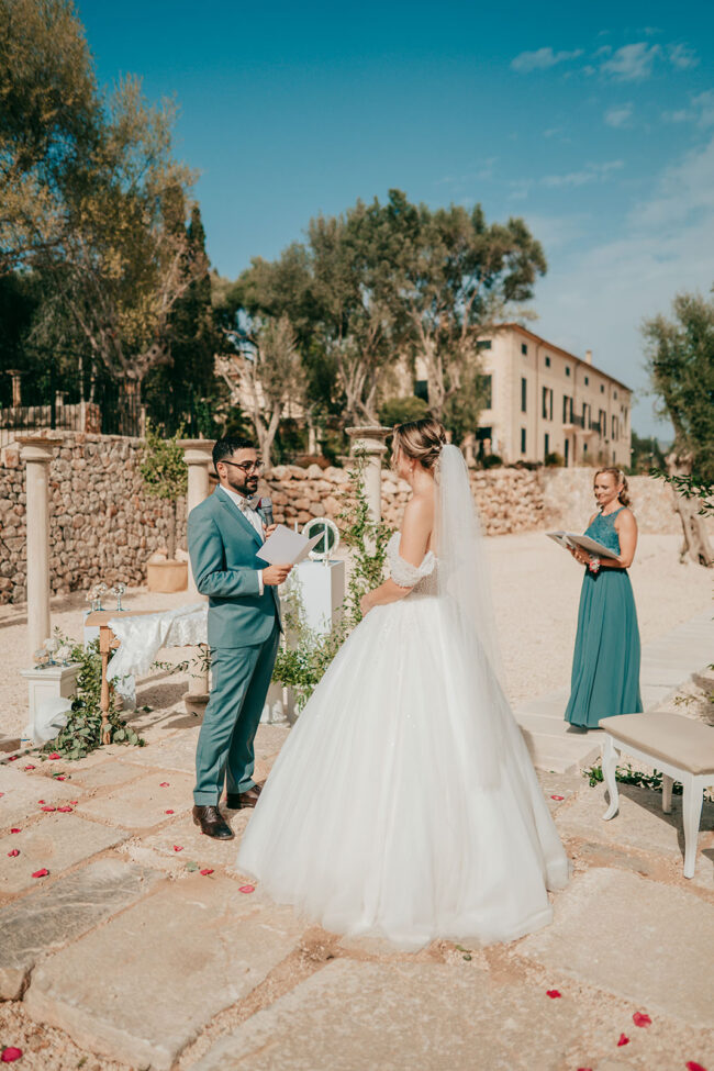 A bride and groom stand facing each other outdoors during their wedding ceremony, holding papers. The bride wears a white gown and veil; the groom wears a blue suit. A bridesmaid in teal stands behind them. The setting is sunny with stone paths.