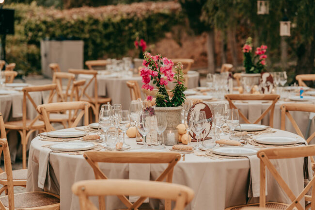 A round table set for an outdoor event with white tablecloth, neatly arranged plates, glasses, and cutlery. A flower centerpiece with pink blooms decorates the table, surrounded by wooden chairs. Other tables are visible in the background.