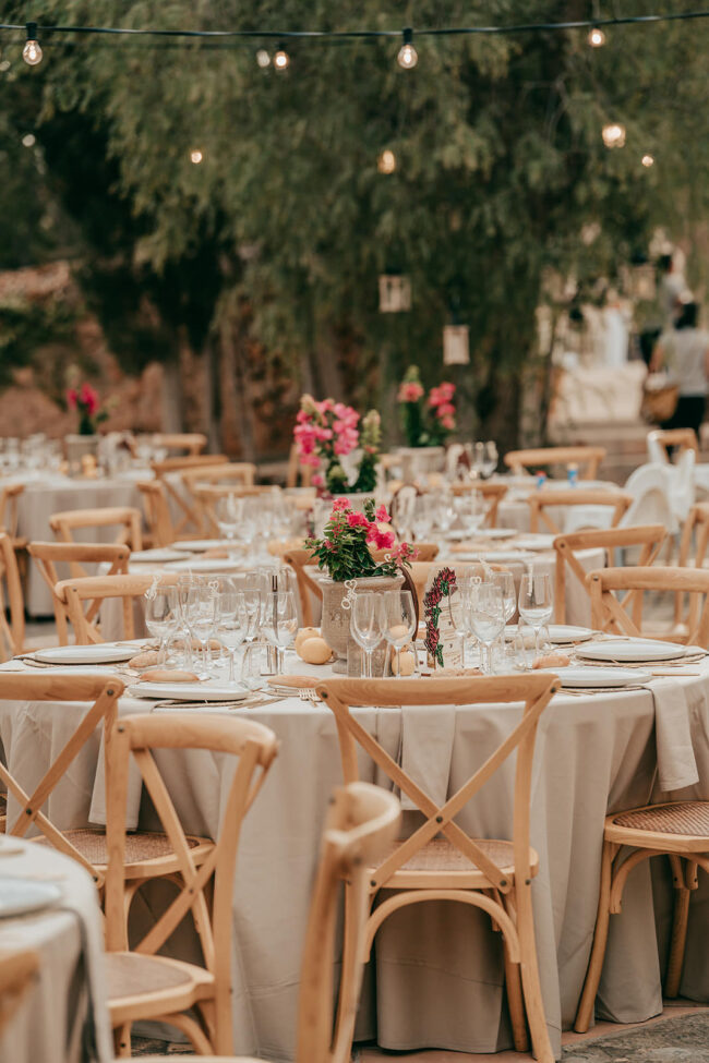 Outdoor event setup with round tables covered in beige tablecloths, wooden chairs, neatly arranged glassware, plates, and floral centerpieces. String lights hang above, and greenery is visible in the background.