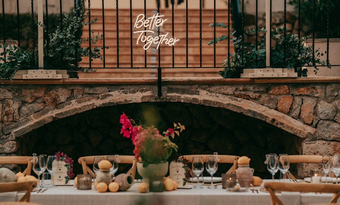 A rustic outdoor dining table is set with glassware, candles, flowers, and lemons. Behind it, a stone wall features a glowing neon sign that says Better Together. Plants and greenery decorate the scene.