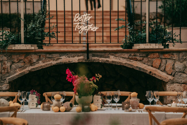 A rustic outdoor dining table is set with glassware, candles, flowers, and lemons. Behind it, a stone wall features a glowing neon sign that says Better Together. Plants and greenery decorate the scene.