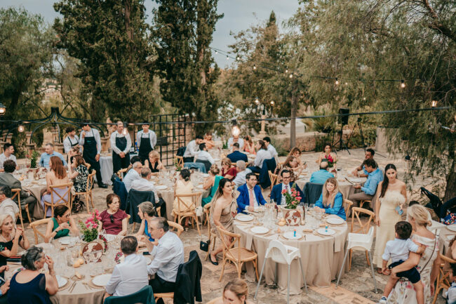 A wedding reception outdoors with guests seated at round tables, decorated with flowers. People are eating, talking, and celebrating. A bride in a white dress walks among the tables, trees and string lights create a festive atmosphere.