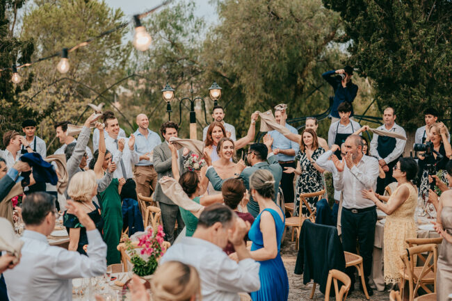 A joyful wedding reception outdoors with guests standing, waving napkins, and smiling as the newlywed couple enters the celebration. String lights hang above, and trees surround the festive scene.