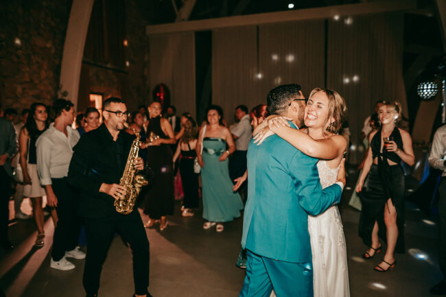A bride and groom joyfully dance together at their wedding reception, surrounded by smiling guests. A saxophonist plays nearby, and warm light shines across the happy crowd celebrating in the background.